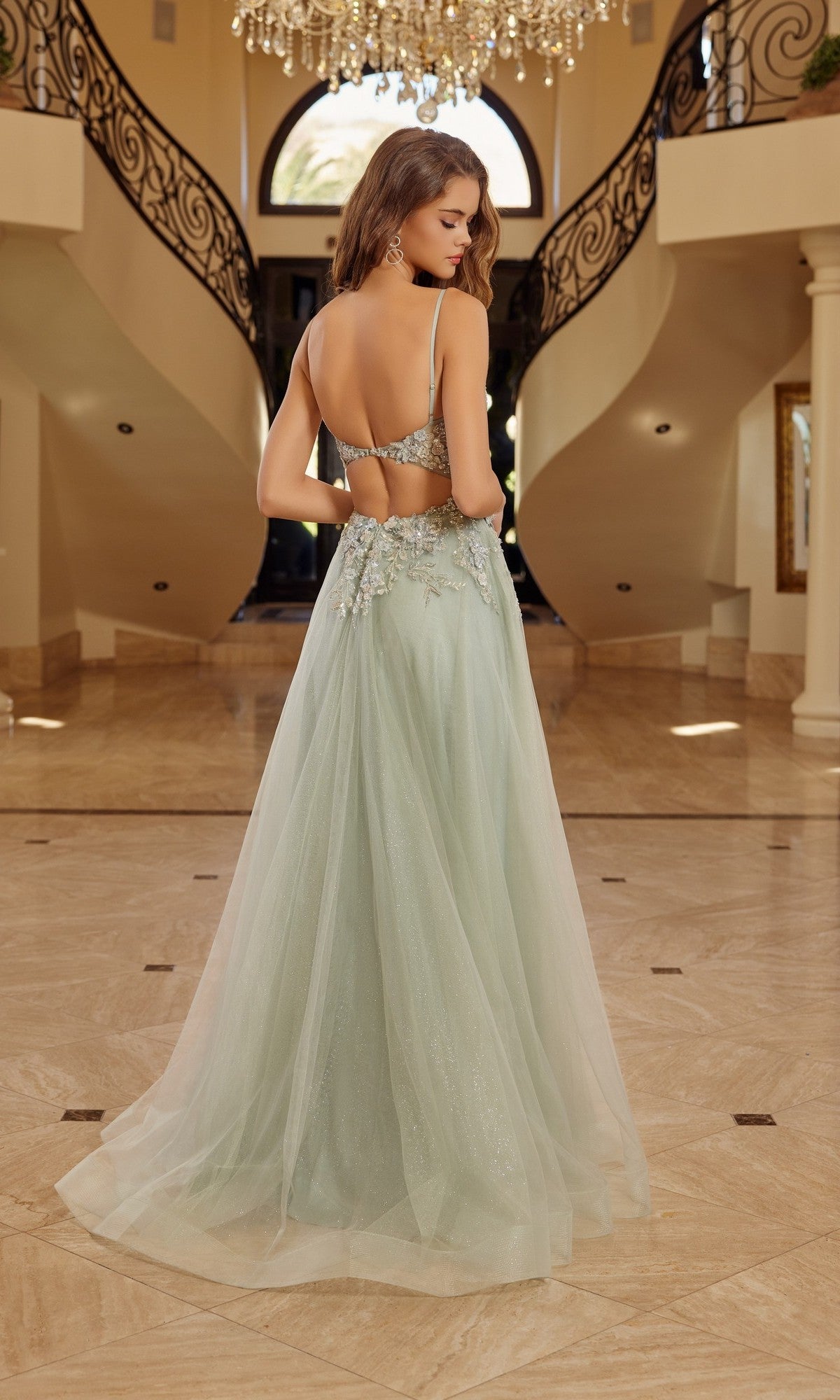 Wearing the Nox Anabel G1787 long prom dress in light green with 3D floral appliques, a woman stands in an elegant foyer featuring marble floors and a grand staircase.